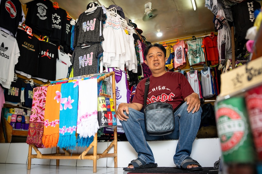 A Balinese man  wearing a red shirt sits on the floor of his shop. The walls are covered in t-shirts and sarongs for sale.