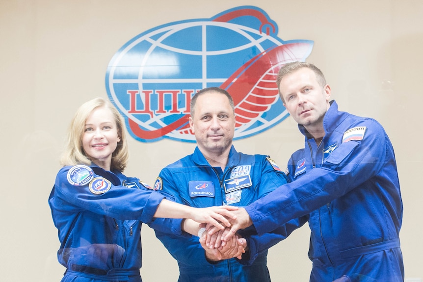 A woman and two men wearing blue cosmonaut suits clasp hands in front of a Russian space agency logo. 