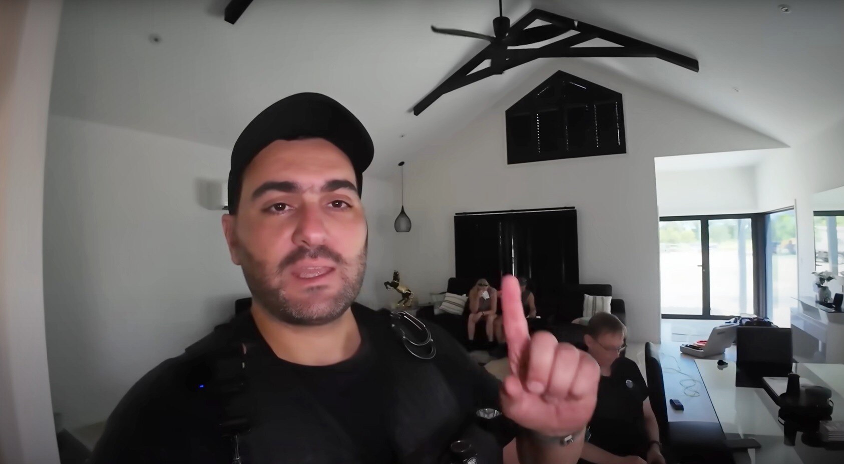 Screenshot from video of a man with cap pointing to roof inside a room