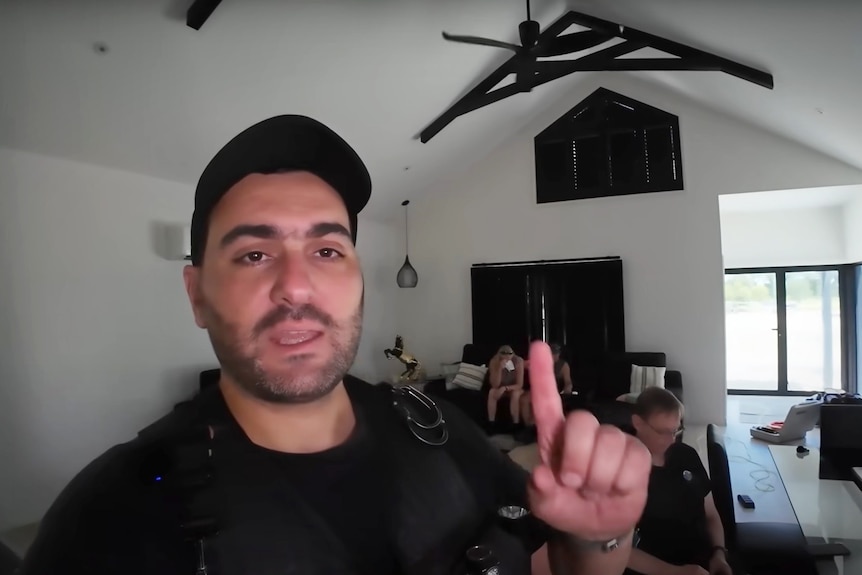 Screenshot from video of a man with cap pointing to roof inside a room