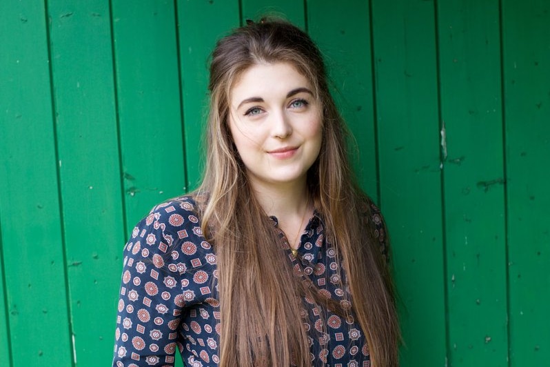 A woman with long blonde-brown hair poses for a photo in front of a green timber wall