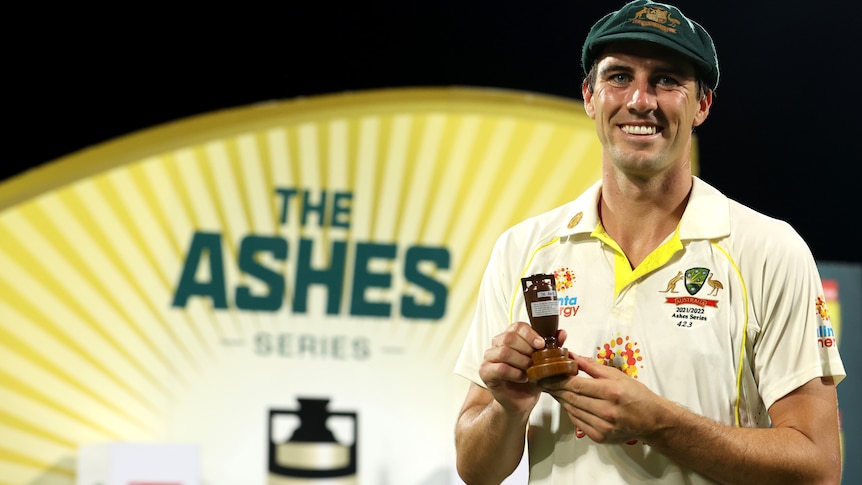 Australian men's cricketer Pat Cummins holds the small Ashes urn in his hands while smiling at the camera