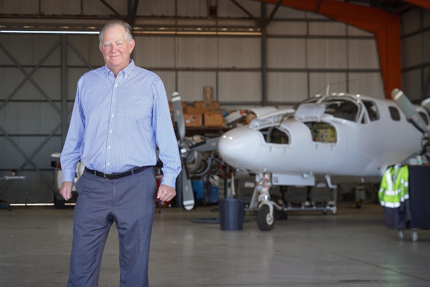 A smiling, older man stands in a hangar in front of a plane.