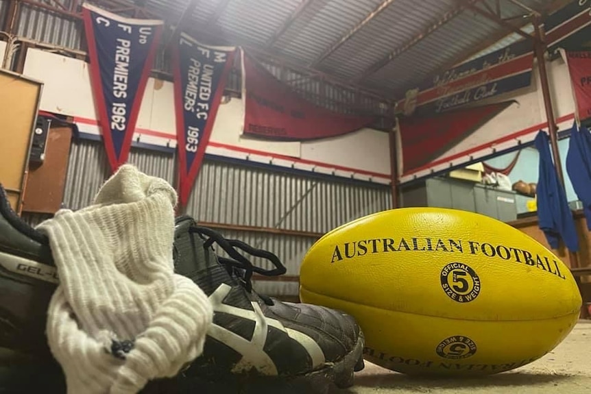 A football boot, a sock and a Australian rules football sit on the ground in front of red and blue banners. 