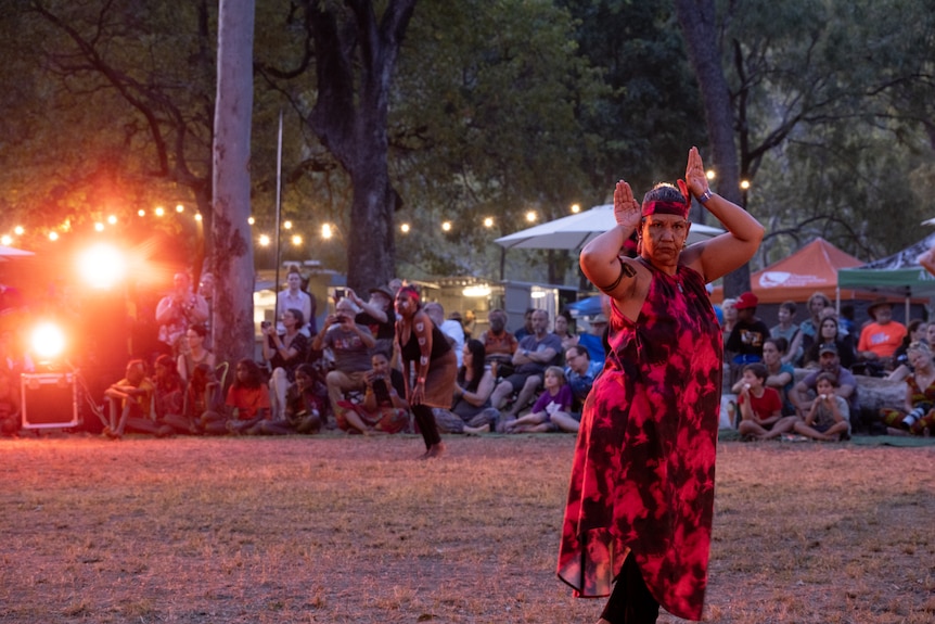 A woman wearing a red and black dress performing outdoors with a crowd and stage lights in the background 