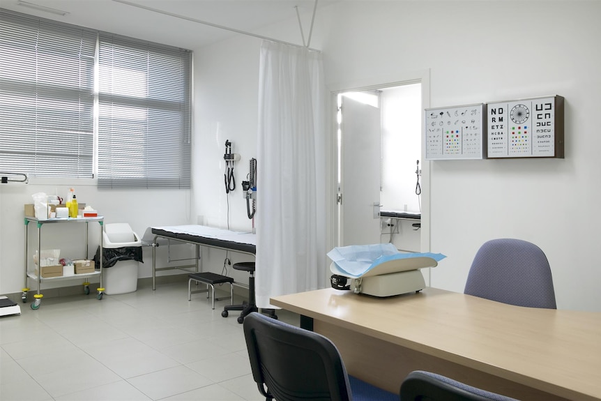 A GP's surgery is seen with a bed for inspection, a caddy with medical equipment, a table, two chairs and an eye test poster.