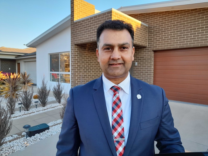 A man in a suit stands in front of a house.