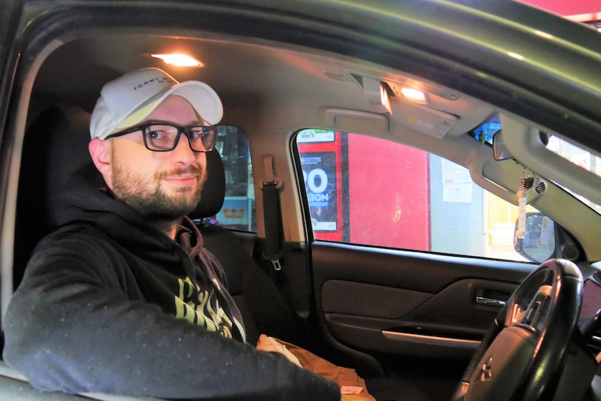 A man with glasses and a cap sits in the drivers seat of his car looking out the window