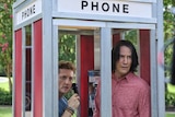 In an outdoor phone box a man with brown hair holds receiver with taller man with dark long hair, both look to outside worried.