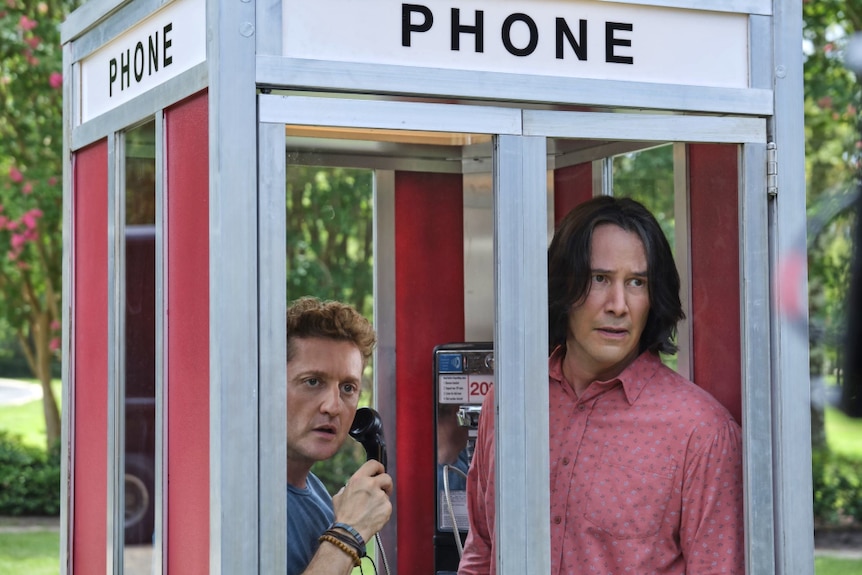 In an outdoor phone box a man with brown hair holds receiver with taller man with dark long hair, both look to outside worried.