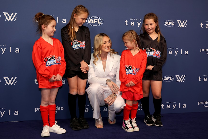 Tayla Harris crouches on the red carpet and is surrounded by four young girls in AusKick uniforms.