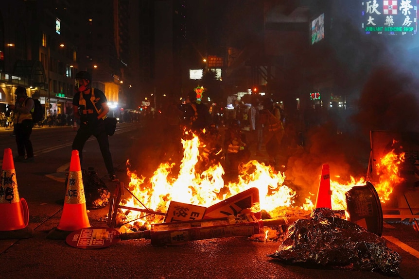 Road signs and traffic cones on fire during night on Hong Kong street with people walking past in background.