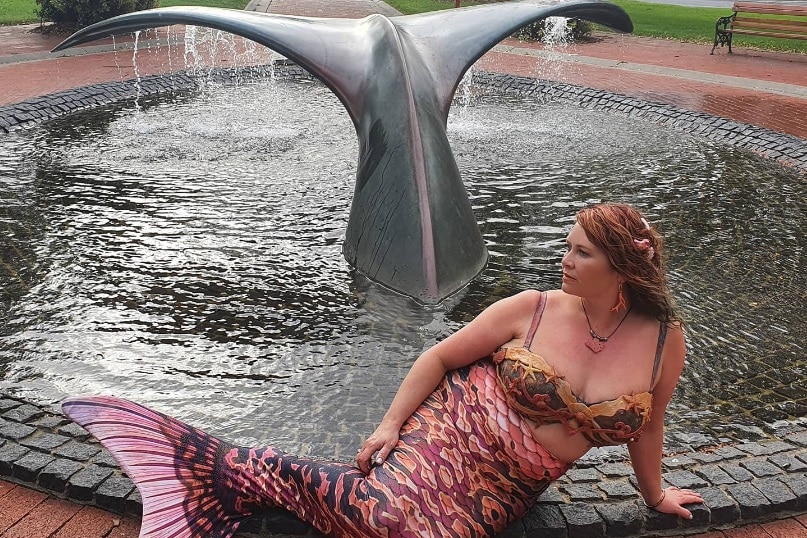 A woman in a mermaid costume poses in front of a whale tail sculpture.