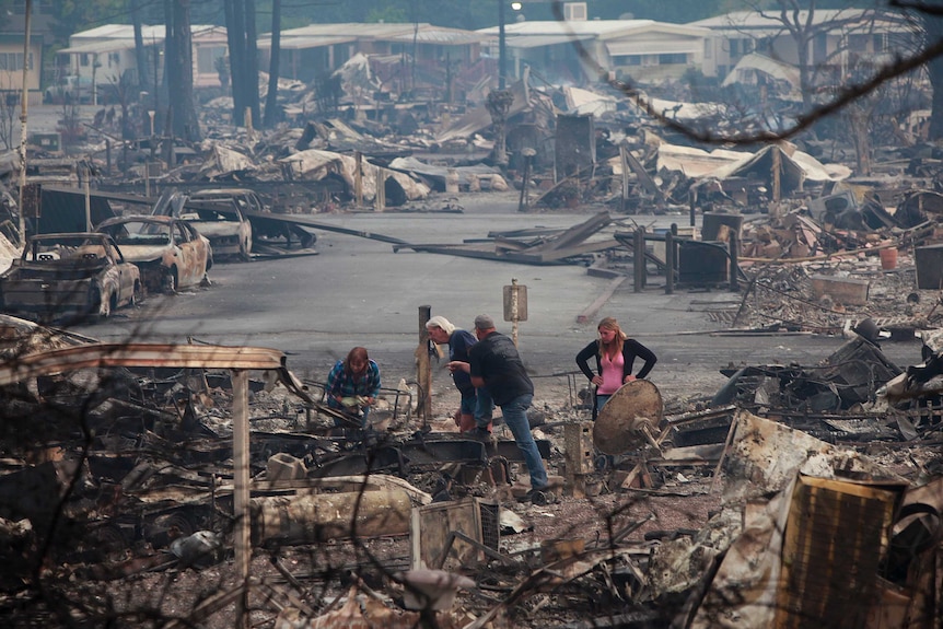 Four people inspect the burned ruins of a house with a whole ruined street behind them.