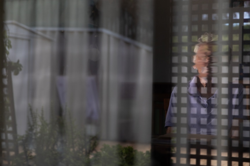 A woman with a blurred face sitting behind a window, the reflection of a lattice looks like prison bars around her.