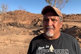 An Indigenous man with a grey goatee stands in the desert, looking serious.