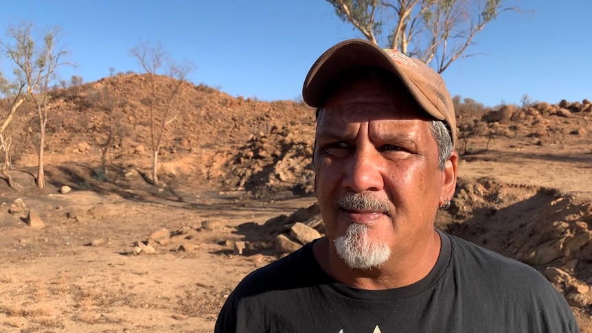 An Indigenous man with a grey goatee stands in the desert, looking serious.