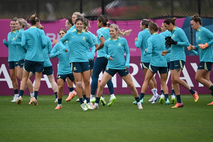 A group of professional female soccer players in teal jerseys running on a pitch in a training session.