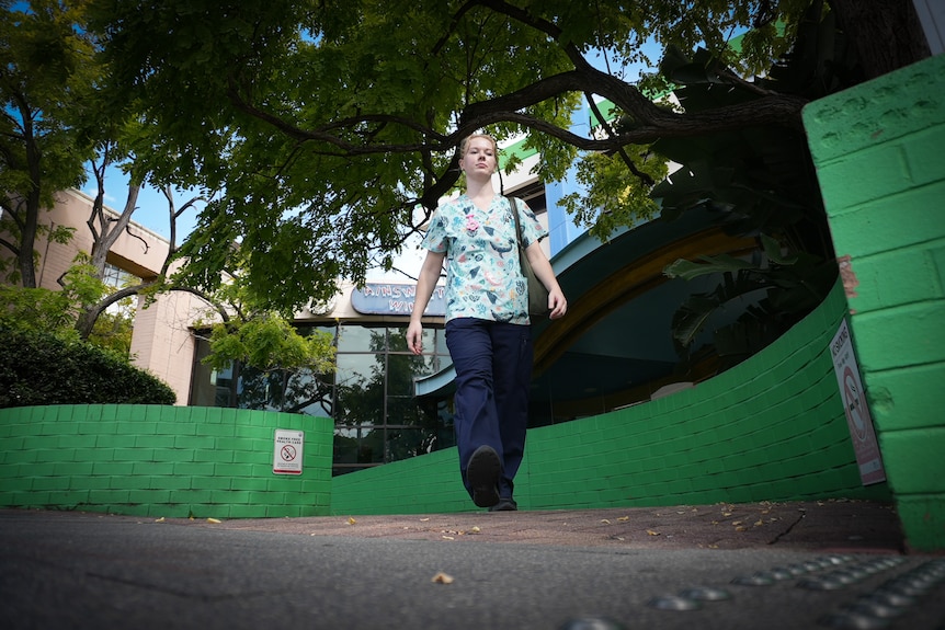 Victoria camina por una calle arbolada frente a un hospital, vestida con una bata médica de colores y pantalones largos.