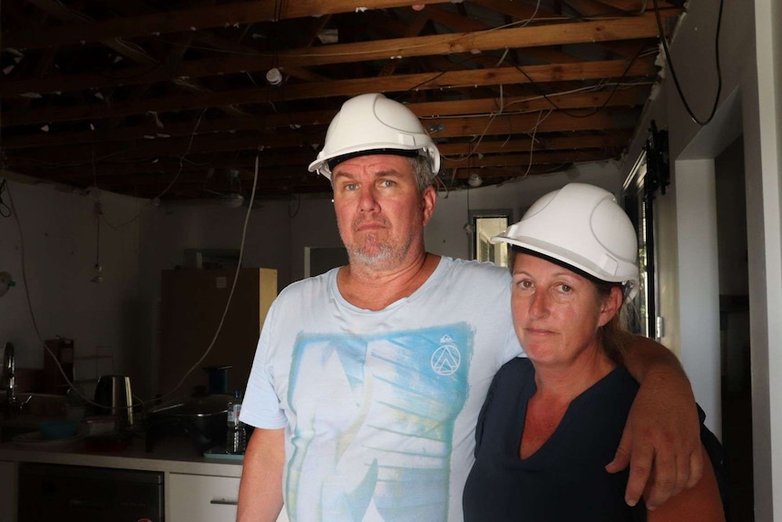 A man with his arm around a woman both are wearing hard hats and are standing in a room where the ceiling has been destroyed