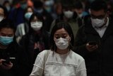 A woman downcast in a white sweater and wearing a mask among a crowd of others at a subway station.