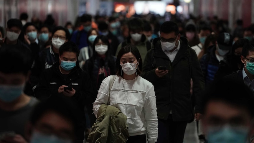 A woman downcast in a white sweater and wearing a mask among a crowd of others at a subway station.