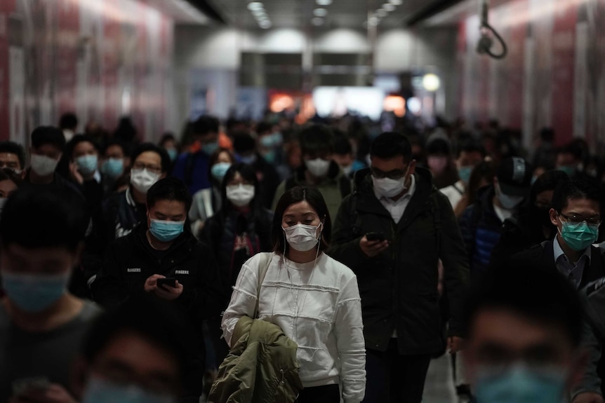 A woman downcast in a white sweater and wearing a mask among a crowd of others at a subway station.