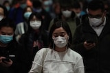 A woman downcast in a white sweater and wearing a mask among a crowd of others at a subway station.