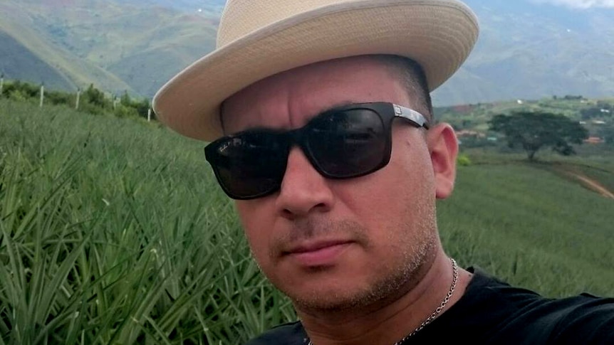 A man wearing a hat and sunglasses takes a selfie against a background of fields.