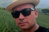 A man wearing a hat and sunglasses takes a selfie against a background of fields.