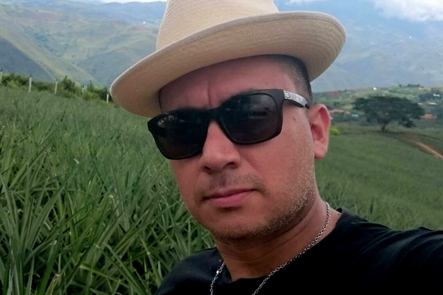 A man wearing a hat and sunglasses takes a selfie against a background of fields.