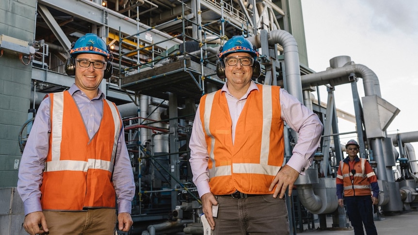 Two men wearing orange vests and blue helments stand in front of a power station