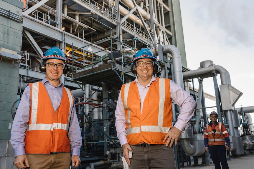 Two men wearing orange vests and blue helments stand in front of a power station