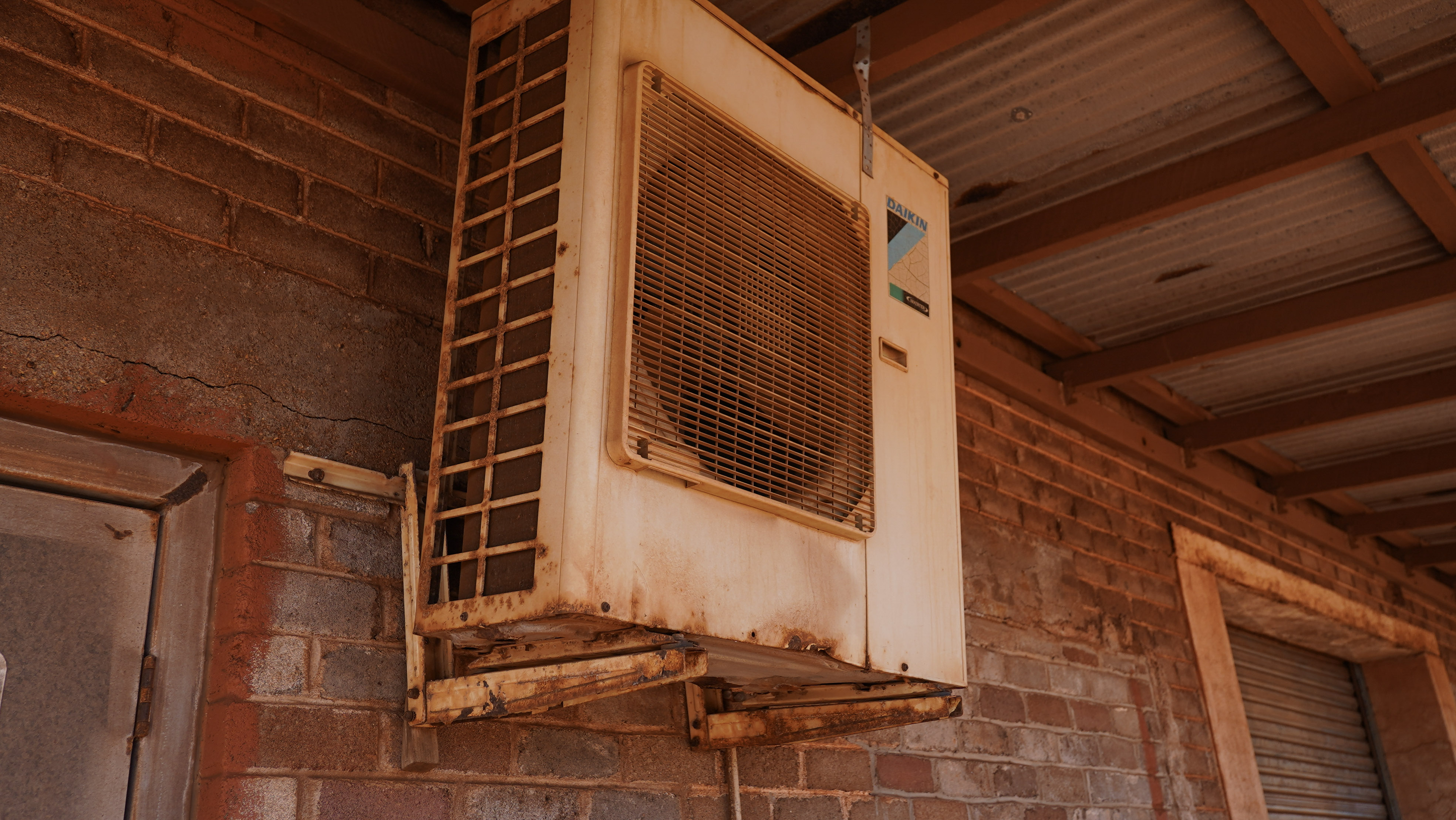 Un aire acondicionado colocado afuera en la pared cerca del techo está oxidado con polvo rojo dentro y alrededor de las aletas.