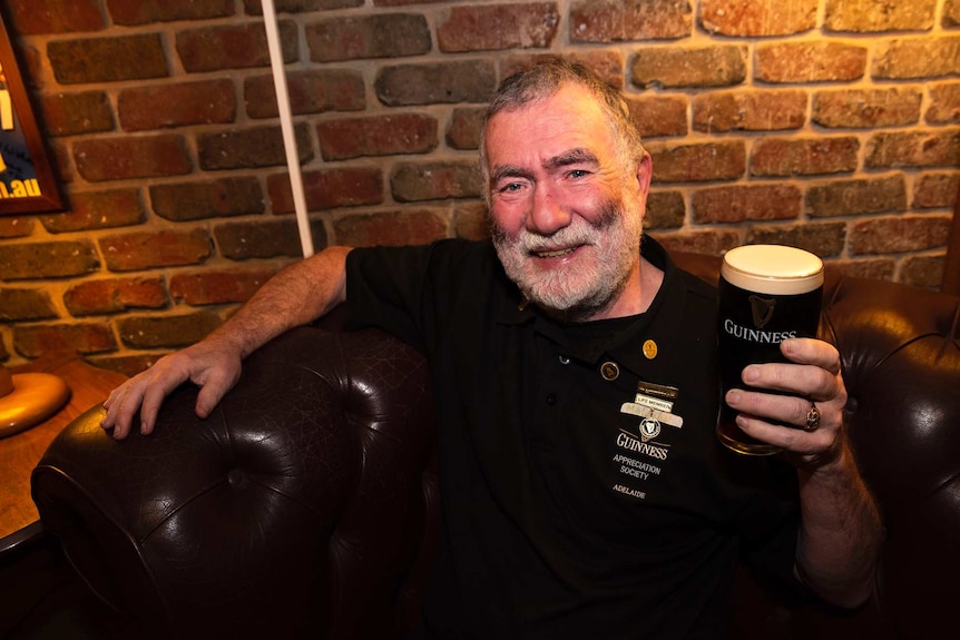 A man sits on a couch holding a full pint of Guinness.