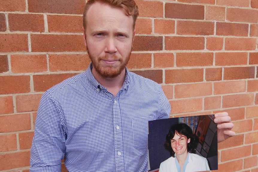 A young man stands in front of a brick wall and holds the photograph of his mother.