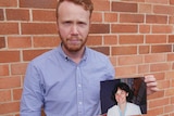 A young man stands in front of a brick wall and holds the photograph of his mother.