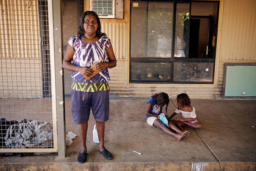 An Indigenous woman stands on her veranda with her two children