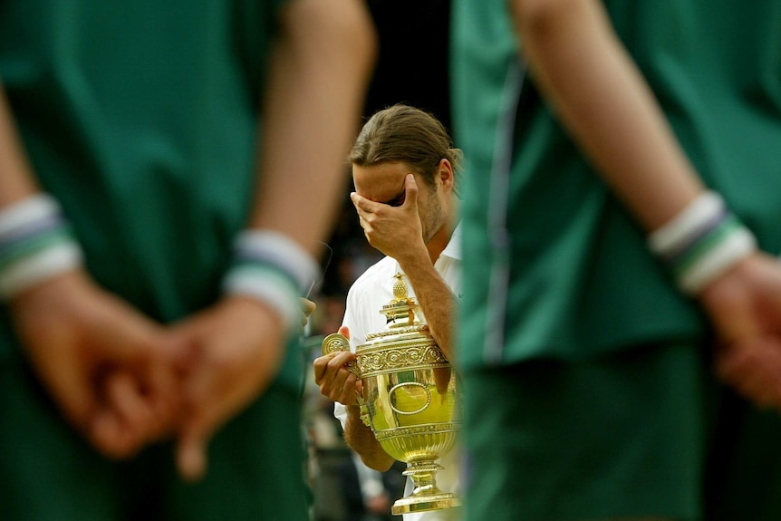 Roger Federer holds the Wimbledon trophy while hiding his face with the other hand, as ballboys stand watch in the foreground