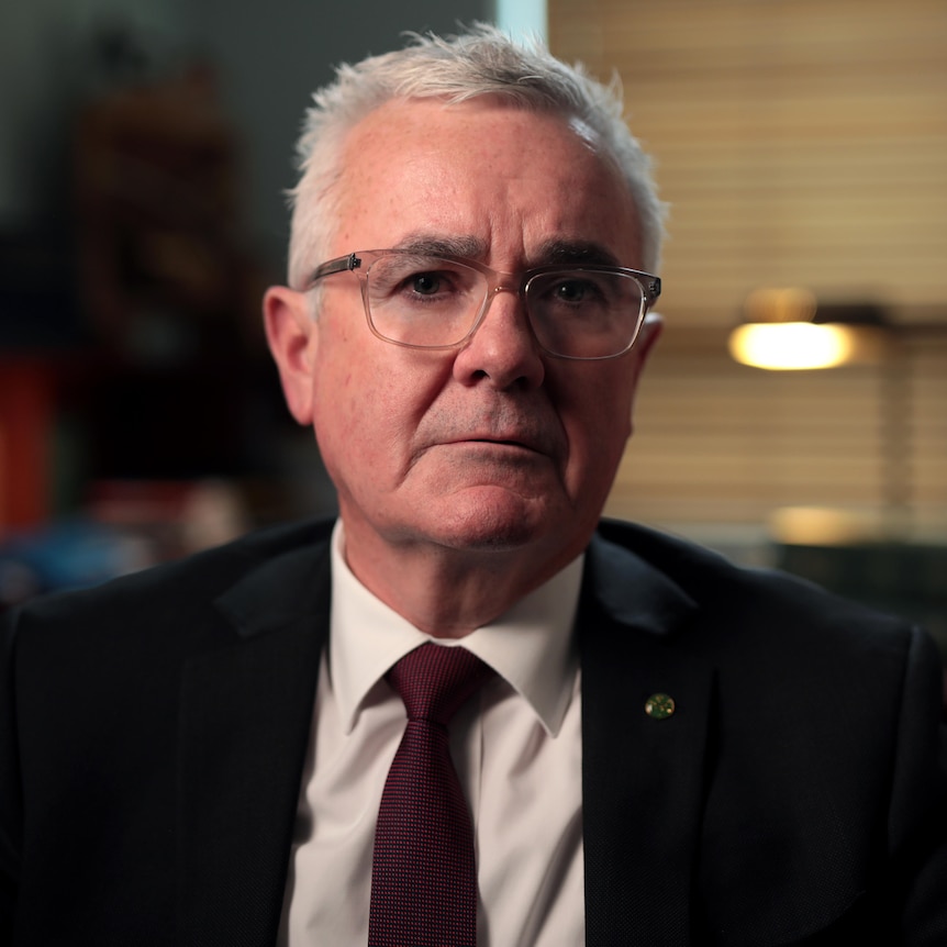 A middle-aged man wearing glasses, a suit and tie, sits in his office looking at the camera. An australian flag is behind him.