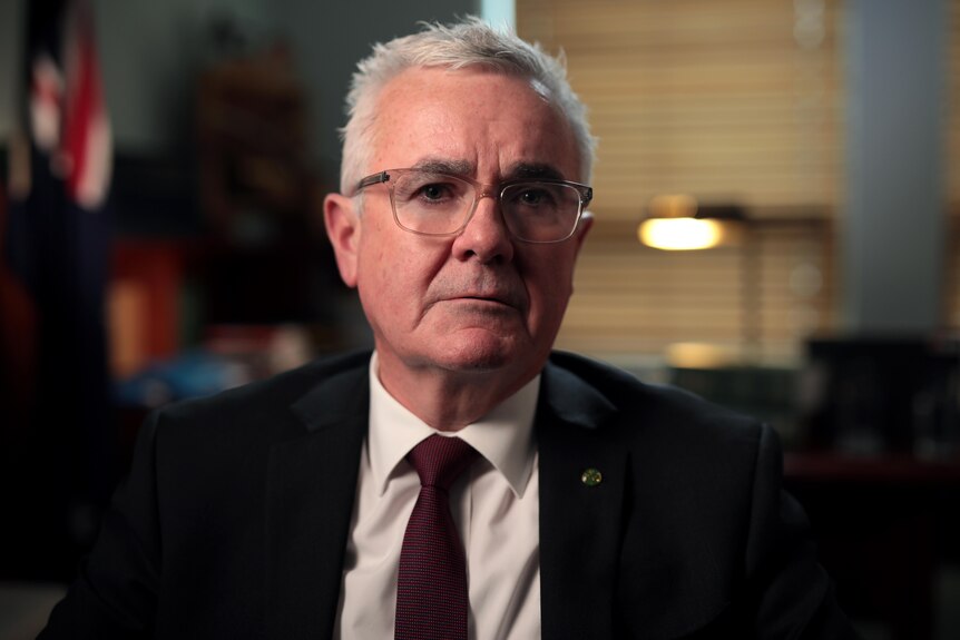 Un hombre de mediana edad con gafas, traje y corbata, está sentado en su oficina mirando a la cámara.  Detrás de él hay una bandera australiana.