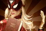 Blonde woman wearing superhero mask and costume, and carrying the Bible.