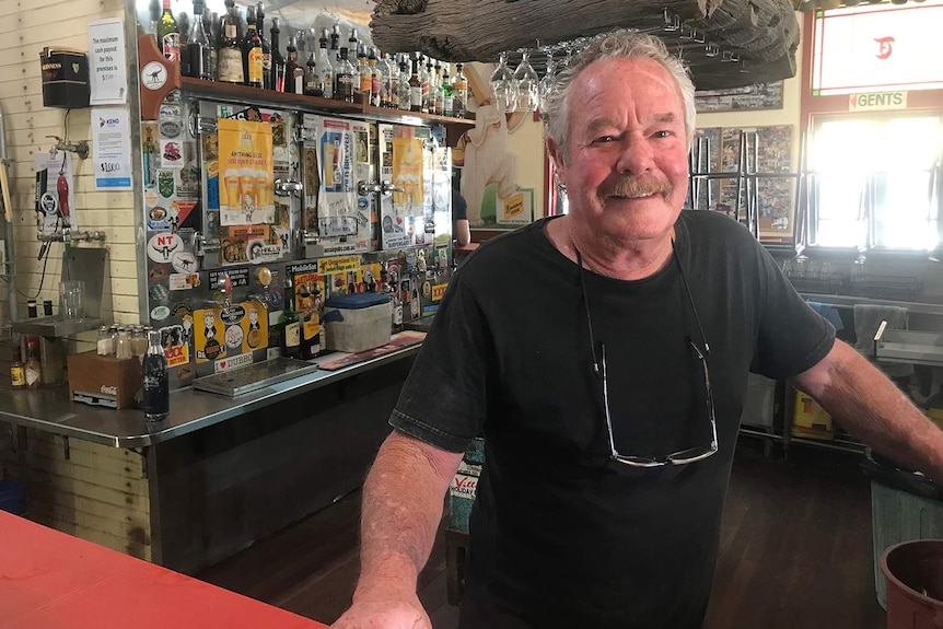 A man in a black t shirt smiles as he stands behind a the bar at an outback pub.