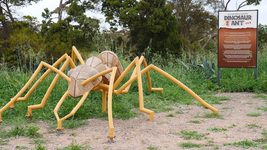 Giant ant sculpture with yellow legs in front of grassy knoll and sigh for Dinosaur ants on right