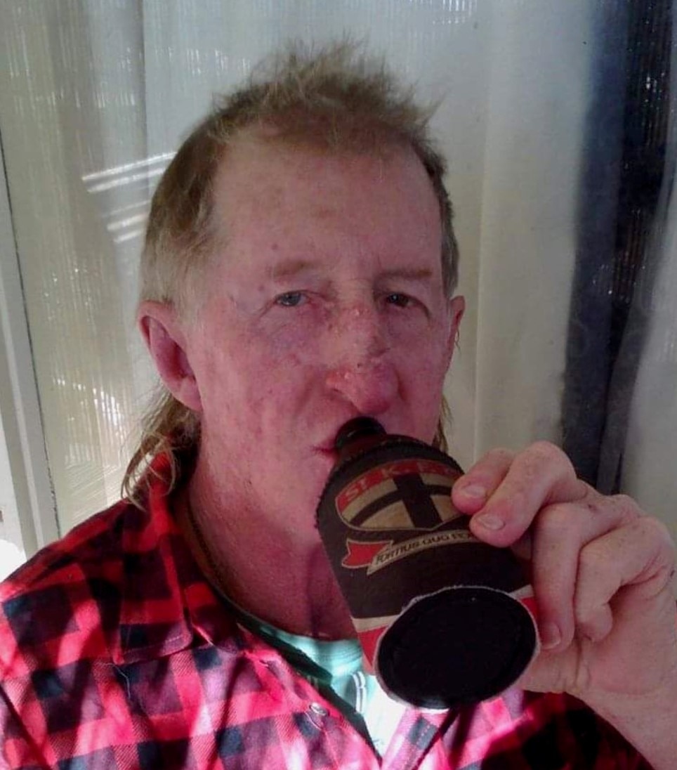 A man wearing a red check flannel shirt drinks a beer from a St Kilda Saints stubby holder