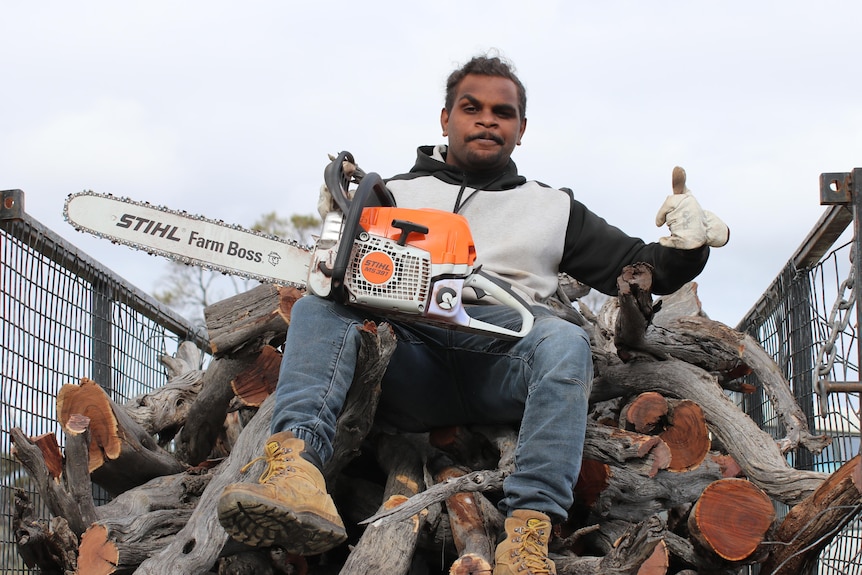 Indigenous man holding chainsaw, sitting up high on pile of wood in high-sided cage trailer.