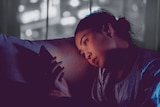 A young woman sits in the dark on the couch, looking at her phone. 