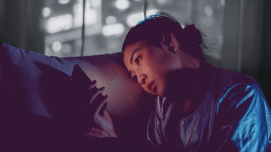 A young woman sits in the dark on the couch, looking at her phone. 