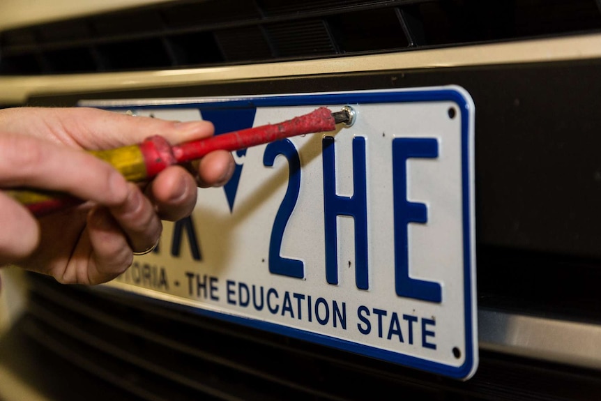 A hand with a red screwdriver trying to unscrew a bolt on a car licence plate.