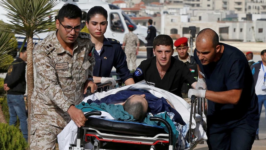 A person on a wheeled hospital stretcher is rushed up a walkway from a helicopter pad, with Anman's skyline viewed behind it.